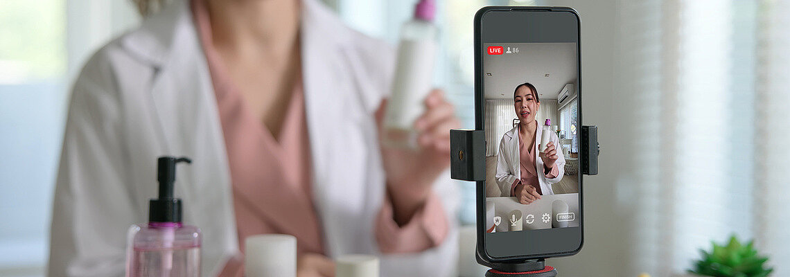 Frau mit weißem Kittel hält verschiedene Produkte vor die Kamera. Aufgestelltes Smartphone filmt.